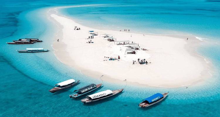 Toma aérea de una pequeña isla de arena blanca en aguas turquesas con botes de madera amarrados y sombrillas.