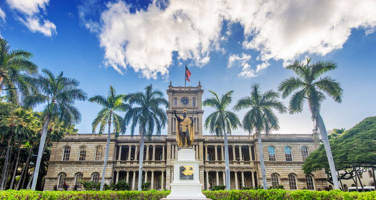 Neoklassiek Iolani Palace en Koning Kamehameha standbeeld omringd door hoge palmbomen onder blauwe hemel.