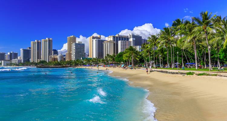 Kristalhelder water en zacht zand van Waikiki Beach met stadsskyline en palmbomen onder een stralende hemel.