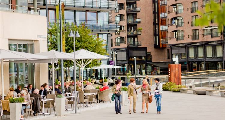 Área de comedor al aire libre con personas sentadas bajo sombrillas, rodeada de edificios modernos.
