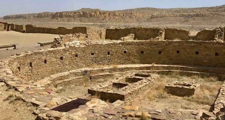Estructura circular de piedra en una zona de ruinas desérticas con acantilados al fondo.