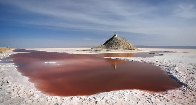 Un paisaje con una colina, un estanque reflectante de agua rosa, y un cielo despejado.