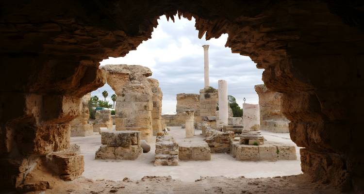 Ruinas antiguas con columnas de piedra vistas a través de una entrada rocosa de cueva bajo un cielo nublado.
