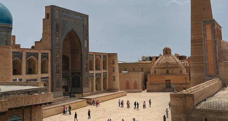 Los viajeros deambulan por un amplio patio rodeado de ornamentados muros de madrasa que presentan un gran portal arqueado y azulejos de mosaico azul.