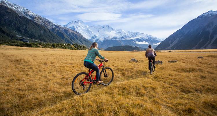 Deux cyclistes roulant à travers un pré alpin doré vers des montagnes enneigées sous un ciel bleu