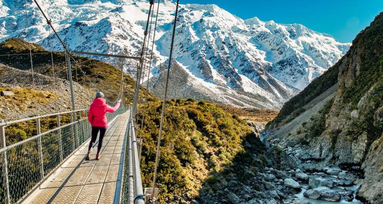 Ein Wanderer in einer rosa Jacke überquert eine Hängebrücke mit hoch aufragenden schneebedeckten Bergen im Hintergrund.