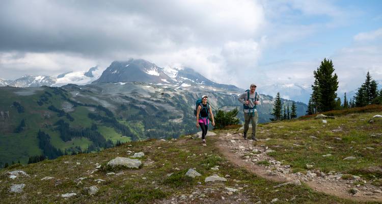 Zwei Rucksacktouristen wandern durch eine Almwiese, während dramatische Gipfel durch aufgerissene Wolken hervorragen.
