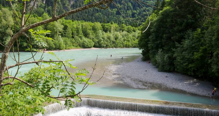Une rivière tranquille avec une petite cascade et un rivage couvert de galets sous un ciel nuageux.