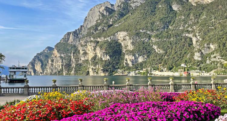 Des parterres de fleurs colorés et une balustrade en pierre encadrent un lac serein avec des falaises majestueuses et un ferry.