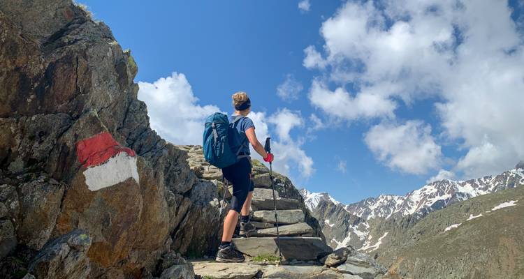 A lone hiker climbing stone steps on a mountain trail with snowy peaks visible.