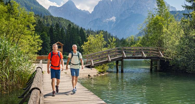 Zwei ältere Männer wandern auf einem Holzsteg über einem See mit Bergen.