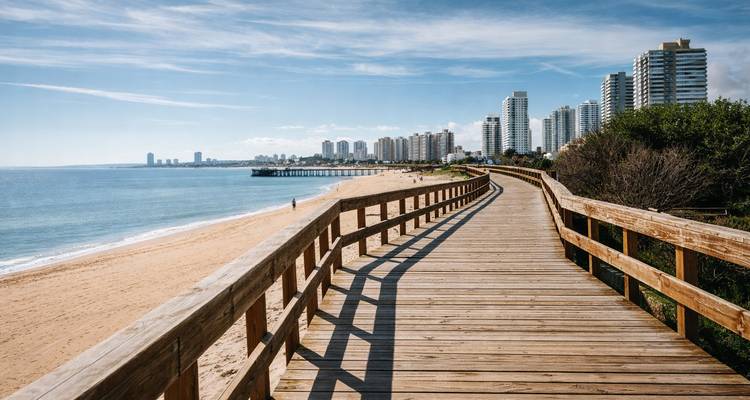 Passerelle en bois le long d'une plage, avec de hauts bâtiments au loin.