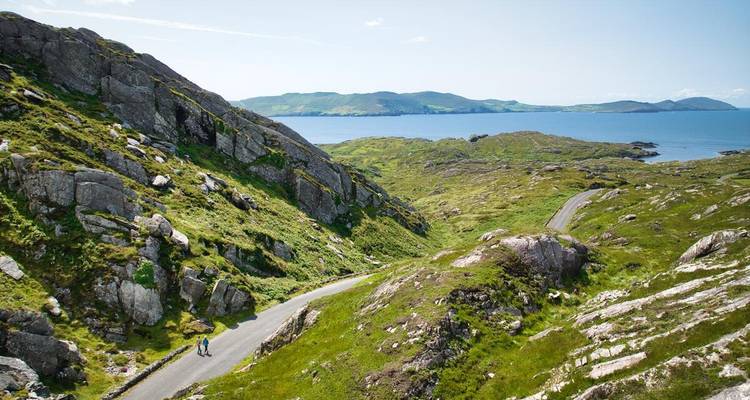 Person walks a winding coastal road through lush rocky hills overlooking the sea