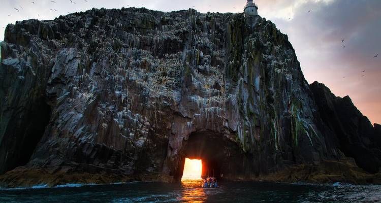 Small boat enters a sea cave pierced by glowing sunset light beneath towering rock cliffs