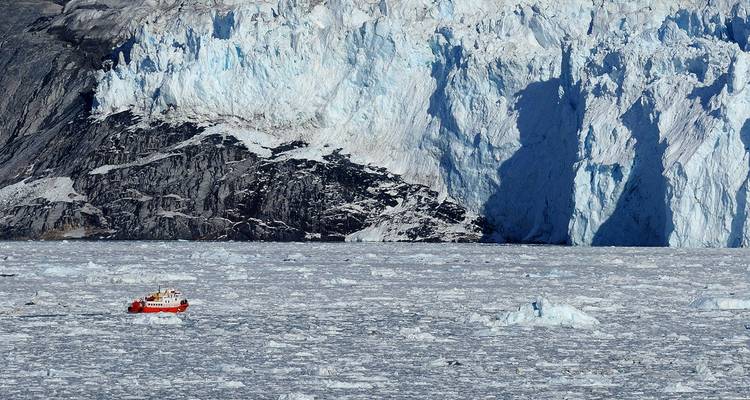 Grand glacier avec des icebergs sur l'eau et un petit bateau rouge au premier plan.