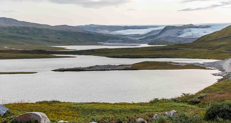 Paysage de toundra s'étendant à perte de vue avec des lacs et des langues de glacier lointaines sous une lumière douce.