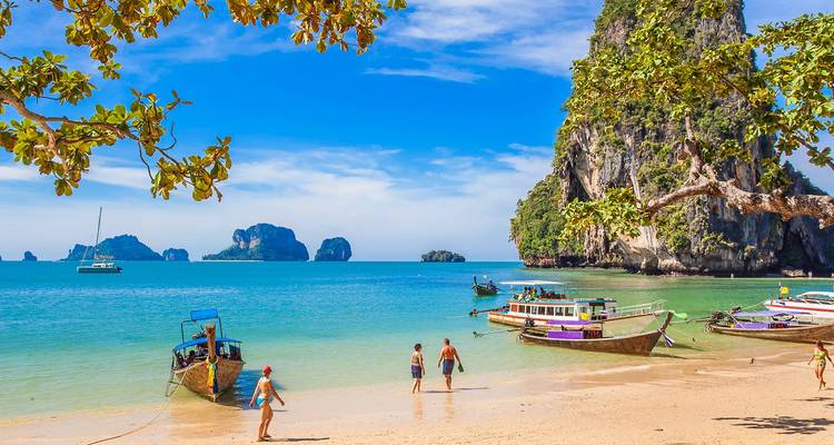 Les visiteurs se promènent sur une plage tropicale avec des bateaux à longue queue et d'imposantes falaises karstiques à proximité.