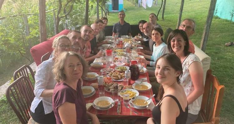 Gran grupo de personas disfrutando de una comida al aire libre, sentadas en una mesa larga rodeada de vegetación.