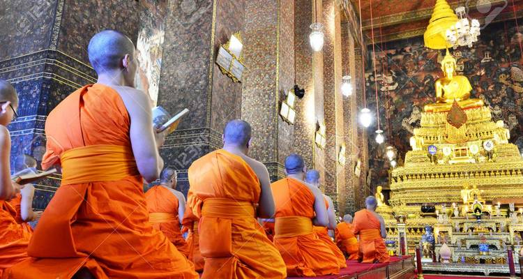 Rows of Buddhist monks in orange robes pray before a golden Buddha statue inside an ornate temple hall.