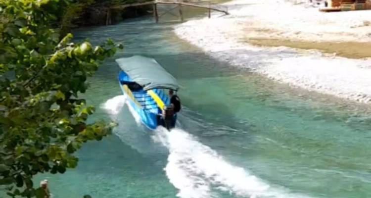 Barco azul navegando por un río turquesa cristalino con orillas de guijarros.
