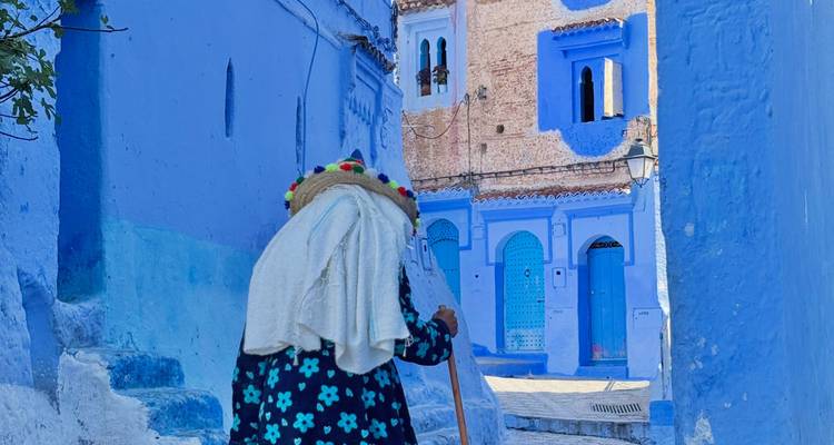 Femme âgée drapée dans un châle blanc et un chapeau de paille montant les marches peintes en bleu dans la médina de Chefchaouen