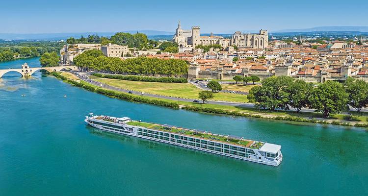 Un élégant bateau de croisière fluviale navigue devant l'horizon historique d'Avignon avec le Palais des Papes et le pont de pierre par une journée ensoleillée.