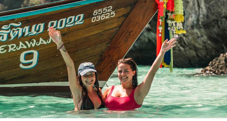 Deux femmes immergées jusqu'à la taille dans une eau turquoise cristalline levant les bras à côté d'un bateau à longue queue décorée.