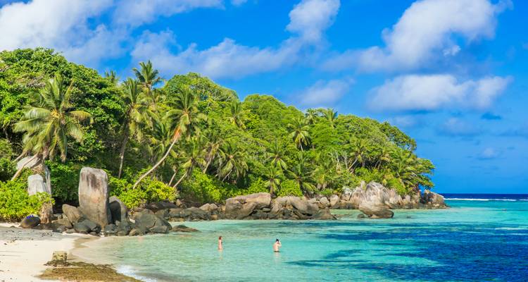 Plage tropicale avec eau turquoise, rochers de granit et forêt luxuriante de palmiers ; deux nageurs dans le lagon