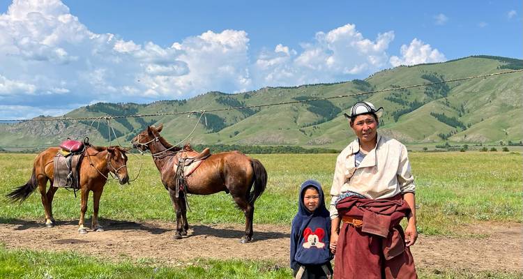 Un cavalier mongol et un enfant posent à côté de chevaux sellés sur une prairie ouverte avec des montagnes en arrière-plan