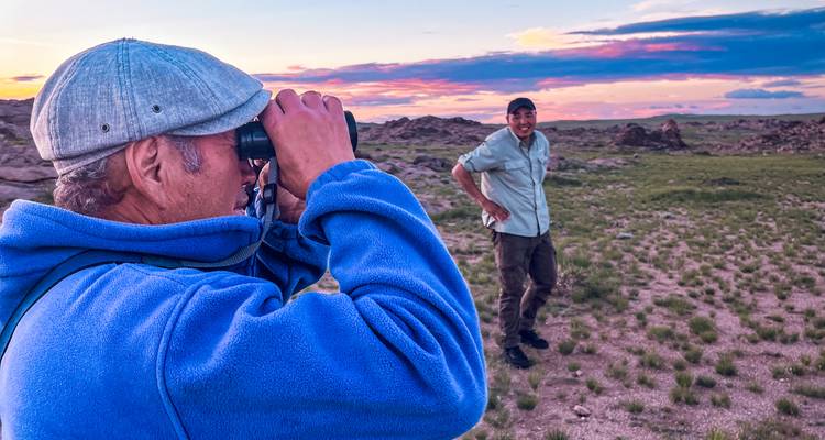 Un voyageur scrute l'horizon avec des jumelles tandis qu'un compagnon sourit pendant le coucher de soleil dans la steppe