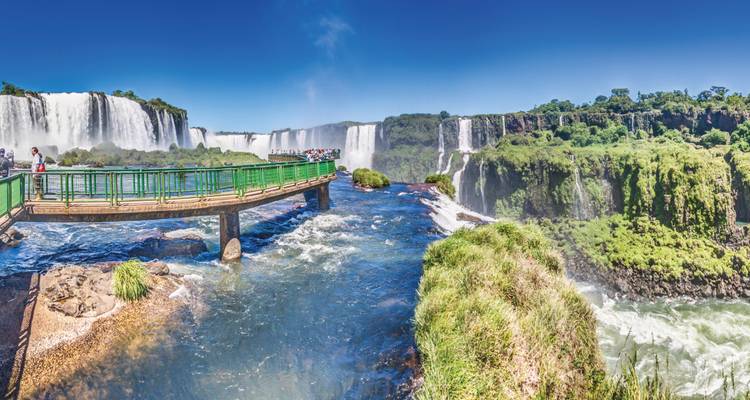 Vista panorámica de las Cataratas del Iguazú con pasarela y visitantes sobre el río caudaloso y la selva exuberante