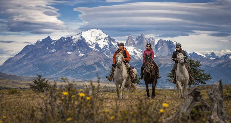 Tres personas montando a caballo frente a montañas bajo un cielo nublado.
