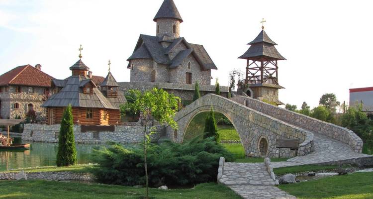 Stone church complex with wooden bell tower and arched footbridge over a pond in a quiet rural setting.