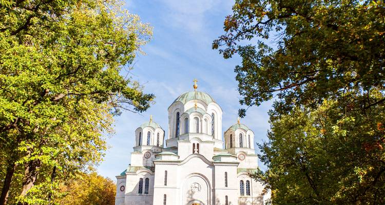 Witte orthodoxe kerk omlijst door groene bomen in een park op een zonnige dag.
