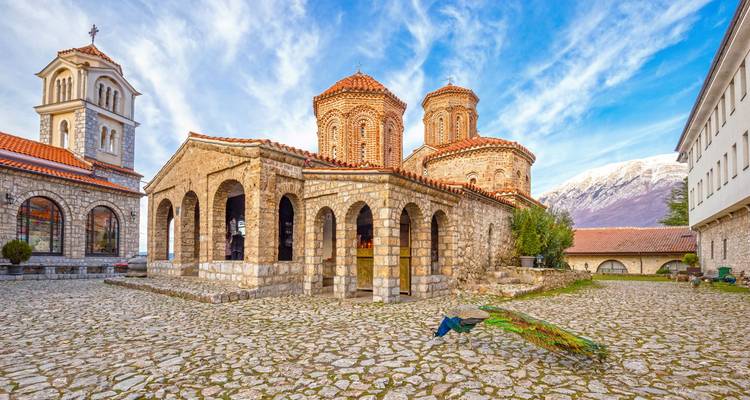 A quaint church with a cobblestone courtyard and mountains in the background.