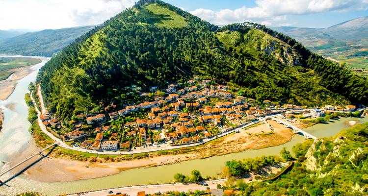 Aerial view of a historic Albanian town wrapped around a lush green hill beside a winding river