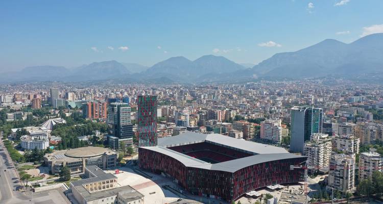 Vista de dron de Tirana mostrando el Estadio Air Albania de color rojo y verde en medio de una vasta expansión urbana y un telón de fondo montañoso.