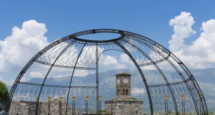 Ruinas abiertas de cúpula de hierro enmarcando la torre del reloj de piedra de Gjirokastra con los Alpes albaneses bajo un cielo azul nítido.