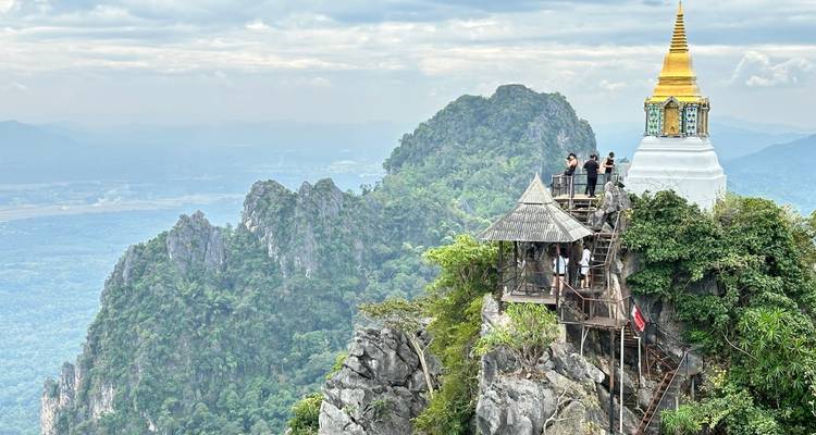 Visitors survey mountain vistas beside a cliffside pagoda reached by wooden stairs.