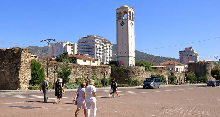Street scene with pedestrians near the historic clock tower and old walls of Elbasan on a bright day.