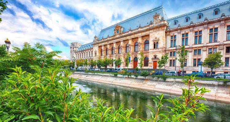 Grand neoclassical Palace of Justice reflected in a canal amid lush greenery in Bucharest.