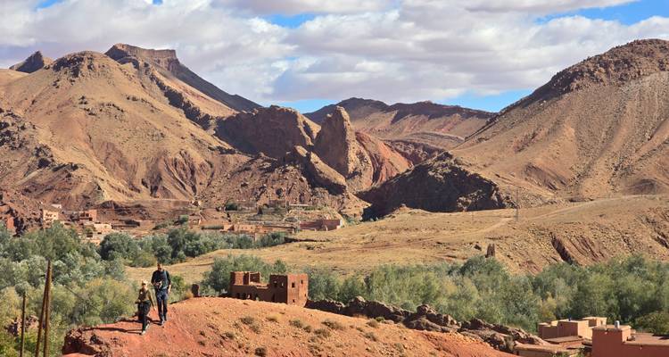 Valle montañoso escarpado con pequeño pueblo y excursionistas en una cresta de tierra roja