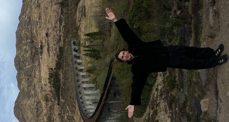 Viajero posa con los brazos extendidos frente al imponente Viaducto de Glenfinnan que se curva a través de las colinas.