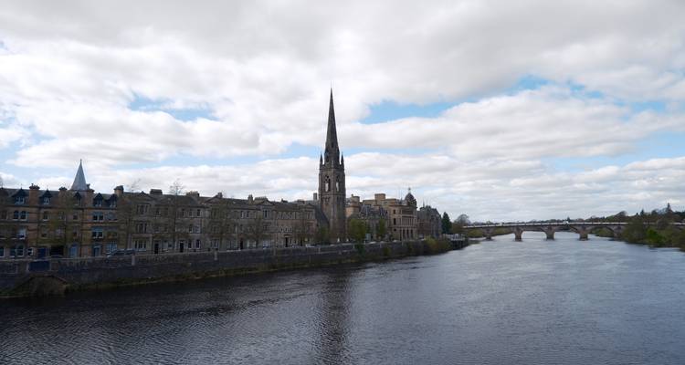 Escena panorámica del río con edificios históricos de piedra y una alta aguja de iglesia reflejándose en el agua tranquila.