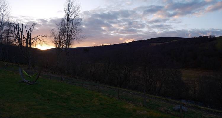 Resplandor del atardecer detrás de colinas onduladas y un valle ligeramente boscoso con nubes dispersas.