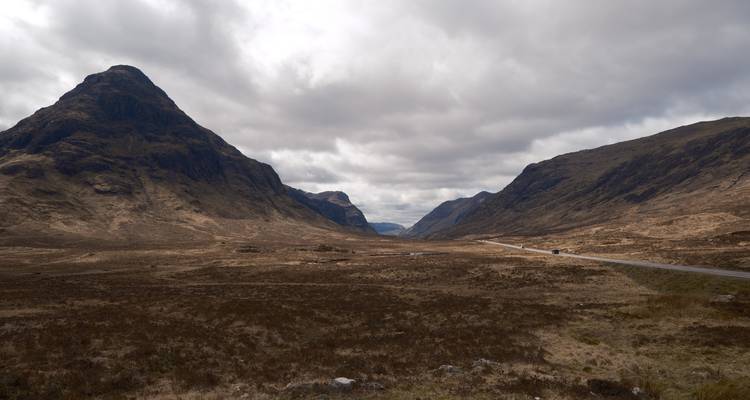 Amplio y escarpado valle de las Tierras Altas flanqueado por montañas empinadas bajo una nubosidad dramática con un camino solitario.