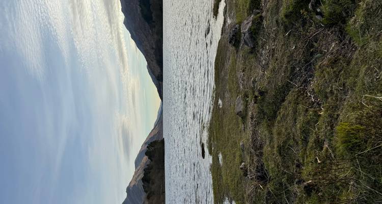 Aguas tranquilas del lago acariciando la orilla cubierta de hierba con colinas arboladas distantes bajo la luz del atardecer.