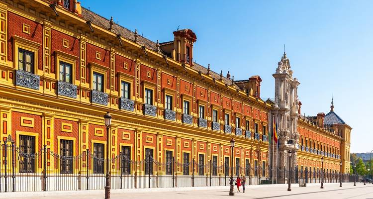 Long ornate Baroque facade of Palacio de San Telmo painted in red and yellow under a clear sky.