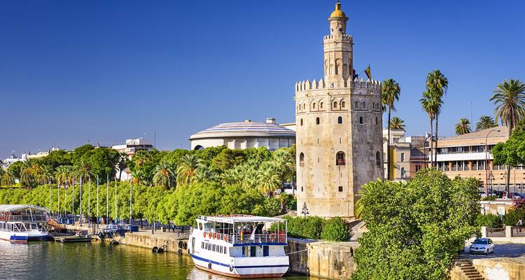 Bright daytime scene of the Torre del Oro on the riverbank with tour boats and palm trees.