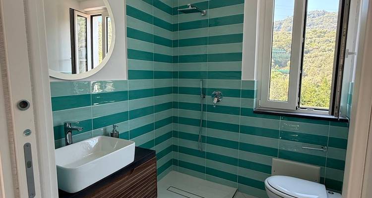 Salle de bain moderne avec carrelage bleu-vert et vue sur les collines par la fenêtre.